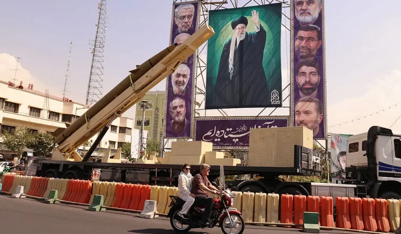 A man rides his motorcycle past a deactivated Kheibar Shekan ballistic missile in front of a picture of Iran's Supreme Leader Ayatollah Ali Khamenei in Tehran's Bahrestan Square on September 27, 2025, as part of an exhibit to mark the "Sacred Defense Week" commemorating the 1980-88 Iran-Iraq war. (Photo by ATTA KENARE / AFP) (Photo by ATTA KENARE/AFP via Getty Images)
